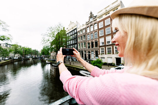 Young happy tourist woman holding up her phone and photographing an Amsterdam canal, Netherlands. Travel and holiday lifestyle concept.