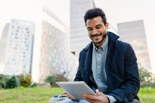 Young adult Latin American businessman using digital tablet at park. Focused professional enjoying a productive outdoor moment. Business and people concept.