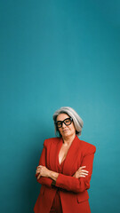 Fototapeta premium Senior businesswoman in a red suit and glasses standing with crossed arms, portraying confidence and empowerment against a blue background