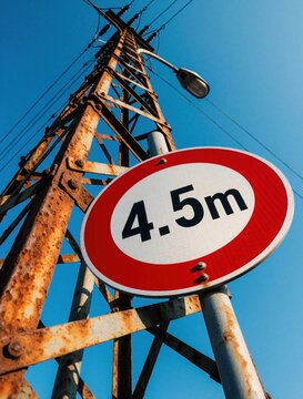 Red traffic sign indicates 4. 5m height limit on rusty metal power pole under clear blue sky. Warning symbol affixed to old industrial pylon. Restricts vehicle passage. Caution sign on weathered
