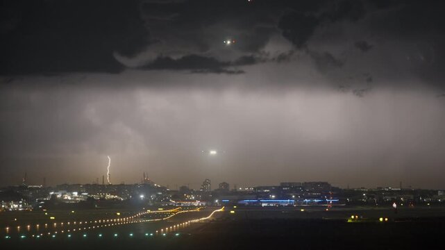 Airplanes queue and approach an illuminated airport runway at night with lightning flashing in the distant sky