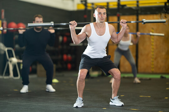 Focused young adult man performing barbell back squats, compound exercise for muscles of hamstrings and glutes during cross training at gym..