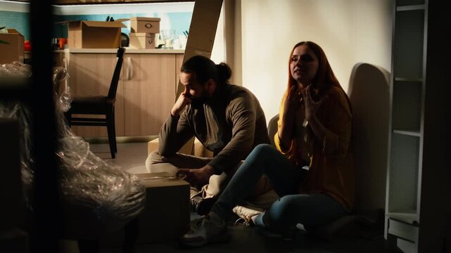 Unhappy partners arguing in a new house among moving boxes and belongings, expressing miscommunication, stress and frustration during relocation and struggling with home organization on moving day.