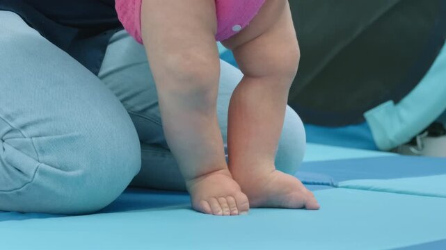 Close-up of a baby's legs and feet standing on a play mat, supported by an adult's hands