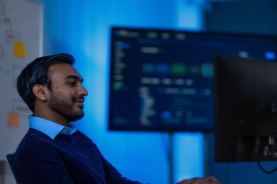 A smartly dressed male engineer in a navy blazer at his monitors watches a chart update while deciding whether to escalate in a modern data operations centre during a busy evening shift