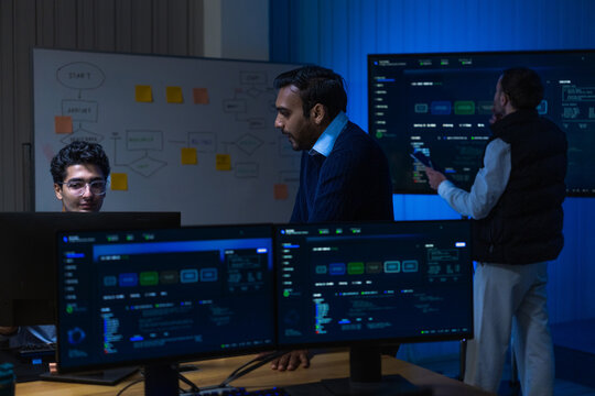 A curly haired male graduate engineer in light blue shirtsleeves lean in together as the metrics finally return to normal in a quiet corporate office in a modern technology company