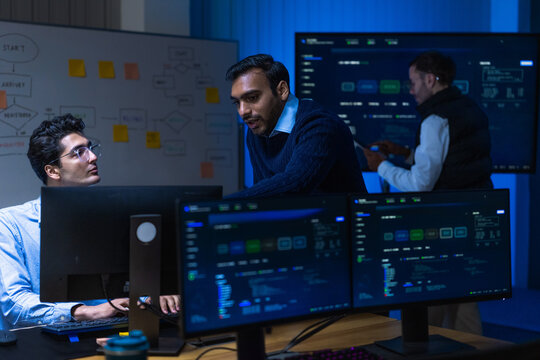 A junior male software engineer in a pale blue shirt holding a digital tablet lean in together as the metrics finally return to normal in a blue lit monitoring room