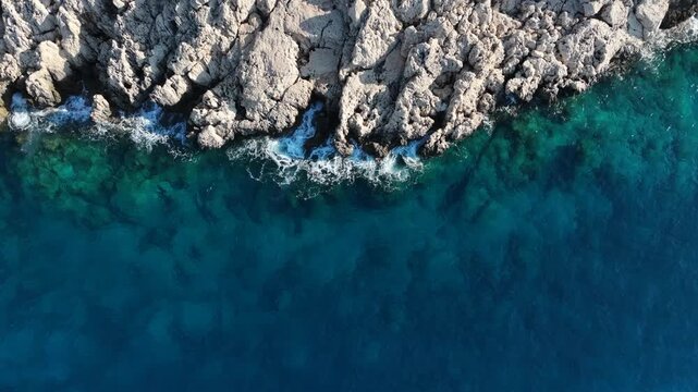 Vertical top-down aerial view of rugged white rocky cliffs and deep blue sea waves crashing on the coast, Antalya region.
