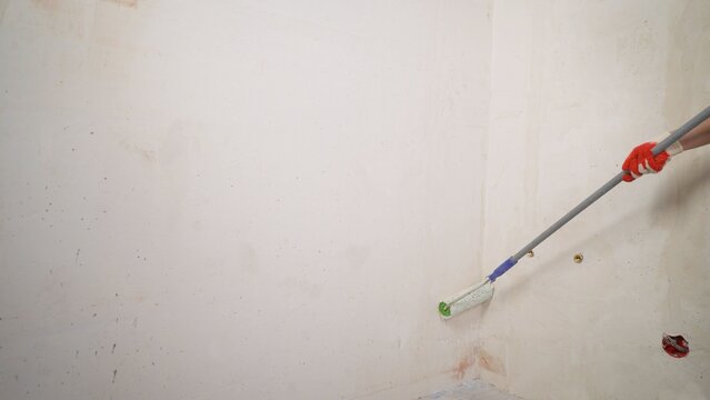 Hand in red glove painting white wall with roller on extension pole, unfinished room corner with exposed fixtures