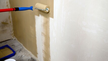 Paint roller applying beige paint to interior wall, blue tray and red handle visible, renovation in progress © Ruslan
