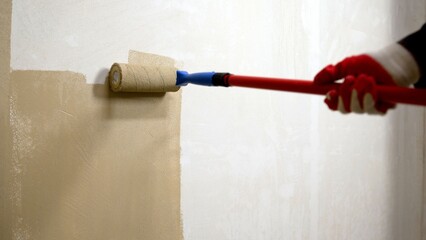 Hand in red glove using paint roller to apply beige paint on white wall, home renovation detail © Ruslan