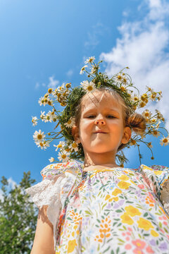 Little girl wearing daisy crown and floral dress, photographed from below against blue sky and clouds. Real childhood, outdoor play, curiosity and connection with nature