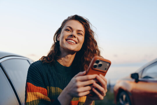 Smiling young woman with curly hair in colorful sweater holding smartphone near parked cars at sunset, joyful moment outdoors, technology and lifestyle concept.