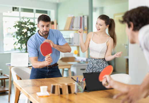 Several office staff are playing game to pass working break time. People play ping pong on working place, they throw ball to each other with help of rackets.