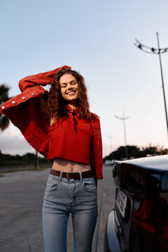 Smiling woman with curly hair wearing a red top and jeans outdoors beside a car. Casual fashion, upbeat mood, soft warm light, and a sense of freedom and confidence.