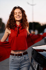 A smiling woman in a red casual top stands outdoors beside a car, arms open. A vibrant daily...