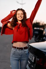 A joyful young woman in a red top and jeans raises her arms and laughs near a car at sunset. Concept of freedom, youth, and carefree happy lifestyle moments. © SHOTPRIME STUDIO