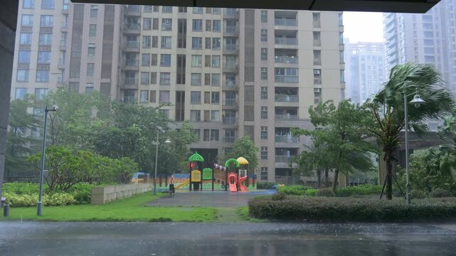 Residential compound playground during heavy wind‑driven rain, wet pavement, swaying palm trees and high‑rise apartment buildings in background video with sound