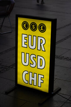 Vertical street city photo. Bright yellow illuminated currency exchange lightbox. Glowing sign with large bold letters EUR, USD, CHF, euro, dollar, pound symbols. Financial theme, business, investing