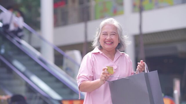 Portrait of Happy Asian active senior woman holding credit card and shopping bag at shopping mall. Elderly retired woman enjoy urban lifestyle shopping with using contactless payment, online banking.