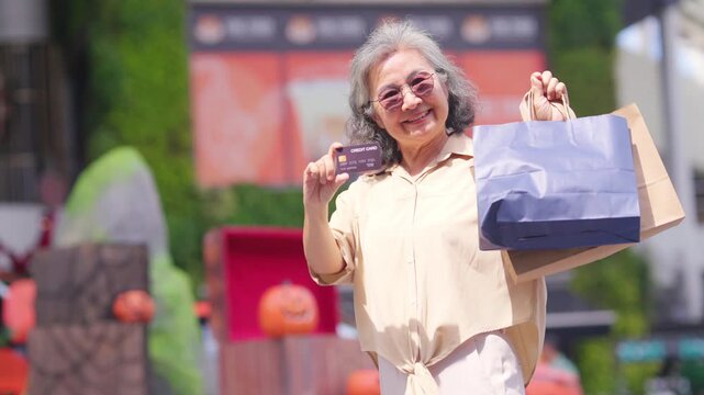 Portrait of Happy Asian active senior woman holding credit card and shopping bag at shopping mall. Elderly retired woman enjoy urban lifestyle shopping with using contactless payment, online banking.