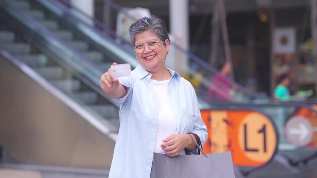 Portrait of Happy Asian active senior woman holding credit card and shopping bag at shopping mall. Elderly retired woman enjoy urban lifestyle shopping with using contactless payment, online banking.