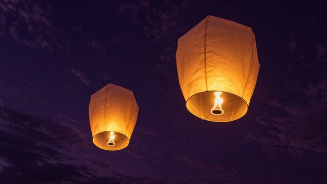 Two illuminated paper sky lanterns float beneath a clouded purple evening sky, with warm golden flames glowing through their textured surfaces.