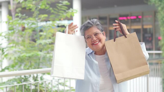 Happy Asian senior woman holding shopping bag at shopping mall, smiling after retail purchase, consumer spending, active retirement lifestyle, independent living, leisure activity and aging gracefully