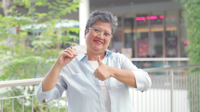 Portrait of Happy Asian active senior woman holding credit card and shopping bag at shopping mall. Elderly retired woman enjoy urban lifestyle shopping with using contactless payment, online banking.