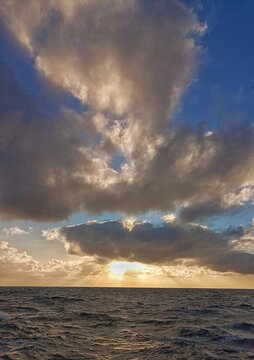 Beautiful vertical seascape of dramatic cloudy sky with warm sun rays breaking through at sunset over dark wavy ocean water