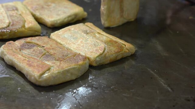 A close-up shot showing the process of cooking martabak telur on a hot flat griddle, resulting in a crispy, golden-brown texture as the traditional dish is prepared.