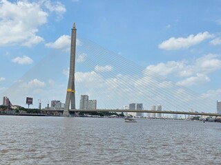 Fototapeta premium Rama VIII Bridge over Chao Phraya River: A grand cable-stayed bridge spans the wide Chao Phraya River, with modern skyscrapers forming the city skyline in the background under a partly cloudy sky.