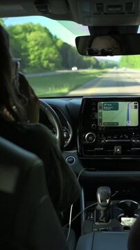 Vertical screen. Female driver with long hair holds a smartphone to her ear while driving on a highway, showcasing a clear view of the road ahead and green trees along the roadside in the background
