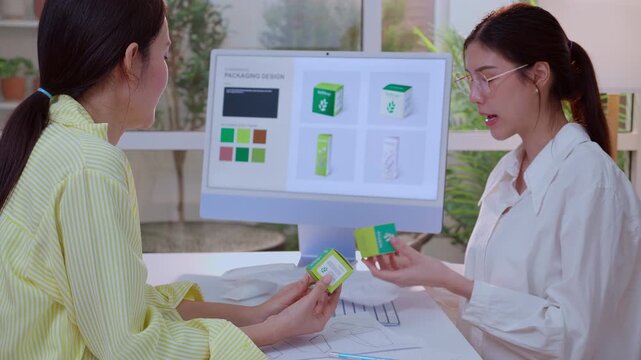 Two asian businesswoman working on packaging design with ai compute and presentation and meeting of cosmetic box mockup product at modern office, planning creative idea of skin care product.