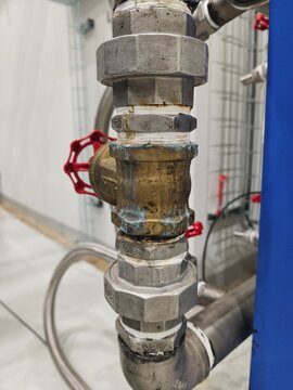 A close-up shot focusing on a leaking pipe joint or brass valve in an industrial plumbing system, showing water droplets or fluid leakage at the connection point requiring technical maintenance 