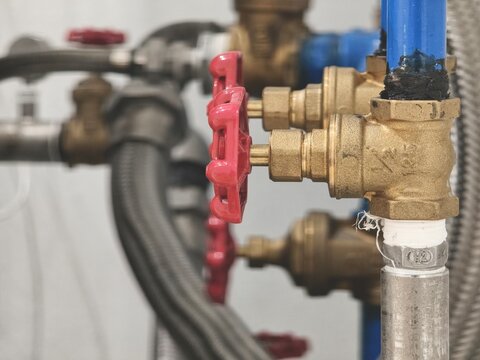 A close-up shot of an intricate industrial plumbing and piping assembly, featuring brass gate valves with red handwheels, blue metal pipes, and flexible stainless steel braided hoses.
