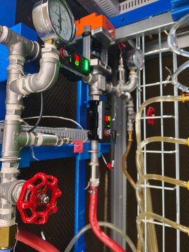 A low-angle shot of a complex industrial plumbing and control setup featuring metallic pressure gauges, a red handwheel gate valve, digital sensors, solenoid valves, and flexible red and clear tubing 
