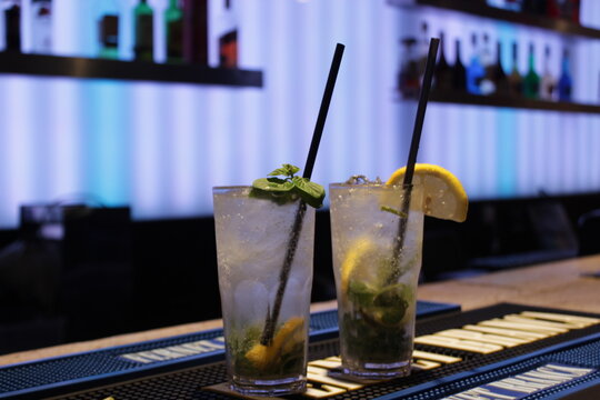 Dos vasos de mojito con hielo, menta y rodajas de lima, sobre la barra de un bar con luces de ne&oacute;n de fondo.
