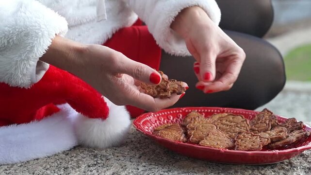 Close-up of a woman in a Santa outfit carefully arranging freshly baked gingerbread cookies on a red festive plate.