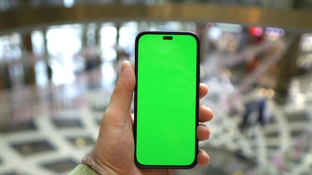 Hand holds a smartphone with a green screen in a mall. The background features blurred escalators and patterned flooring. The phone is viewed closely in the second keyframe