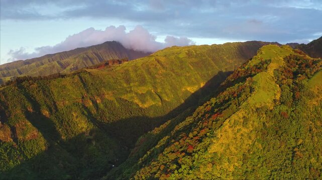 Amazing drone flight over sunset Moorea island landscape with rugged, jungle covered mountain hills in warm sunlight, dramatic shadows casting over valleys and hints of clouds clinging to the peaks