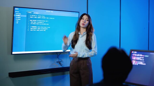 Confident Asian female software developer gives an engaging coding presentation, pointing to a large digital screen displaying computer scripts to a colleague in a modern dark blue tech office.