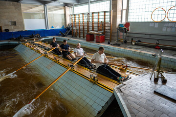Four male rowers train together on fixed machines beside indoor pool. Coordinated crew workout develops rhythm, strength, endurance, teamwork and competition readiness for races. © DimaBerlin