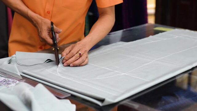 Man worker at clothing factory preparing textile for sewing, cutting fabric cloth with pants pattern using scissors at worktable in workshop, professional employee works at local factory in Vietnam.