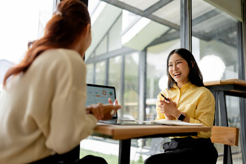 Fototapeta premium Two cheerful Asian businesswomen discussing work and sharing ideas during a casual meeting in a bright workspace
