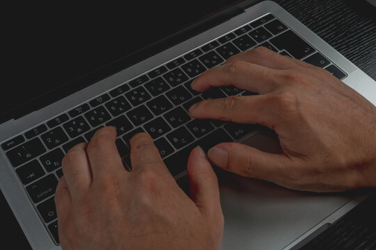 Close-up view of a man's hands typing on a laptop with a Japanese keyboard featuring hiragana characters, in low ambient light.