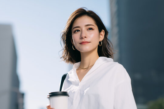 Young Japanese woman wearing white shirt and holding takeaway coffee cup outdoors, confident expression, urban background, lifestyle portrait in natural daylight