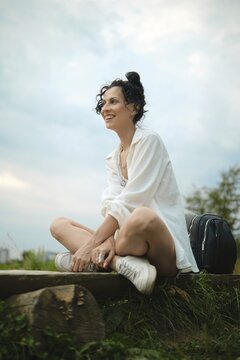 Laughing woman sits on wooden log in grassy place with backpack by her side. She smiles while looking at open space. Sky is cloudy and creates a soft atmosphere around her