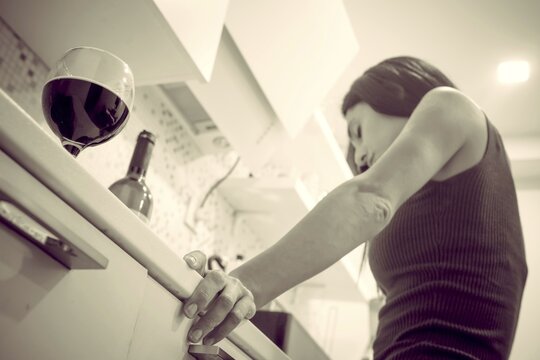 Young attractive caucasian woman hodls hand on kicthen table and feels bad with bottle of wine standing on shelf. Balkans. Croatia