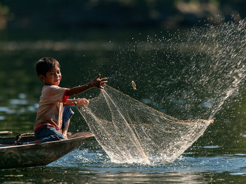 Young boy casting a fishing net from a small wooden boat on a calm lake. Sunlight catching water splashes during traditional fishing.